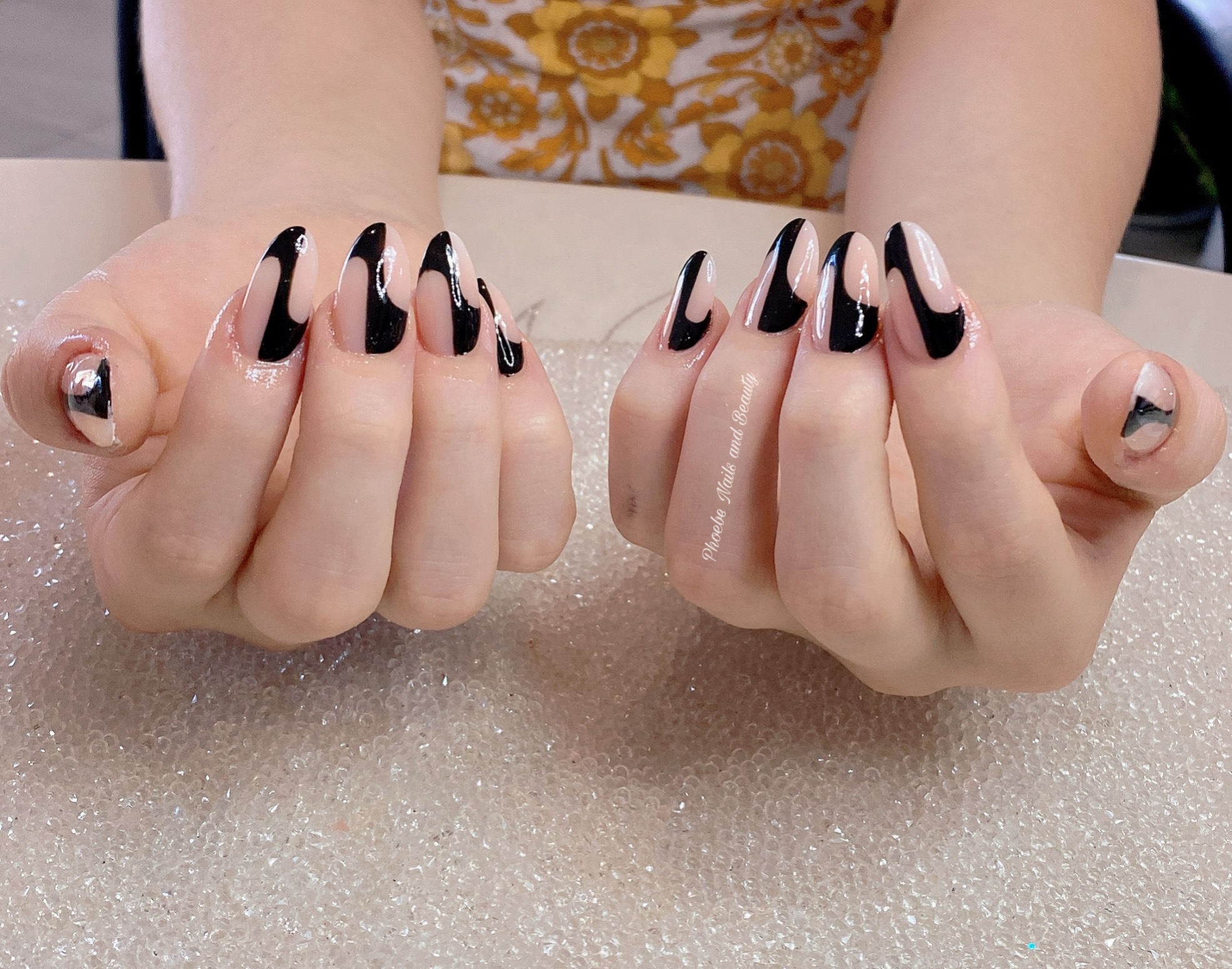 Nude almond nails with glossy black swirl French tips resting on a sparkle mat